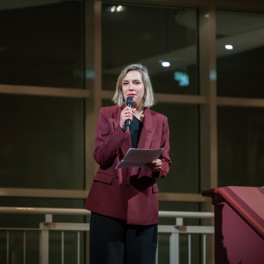 Festival opening in the foyer of the Schauspielhaus Hannover. The photo shows Melanie Zimmermann giving the opening speech. She stands on a staircase and speaks into the microphone in her hand.