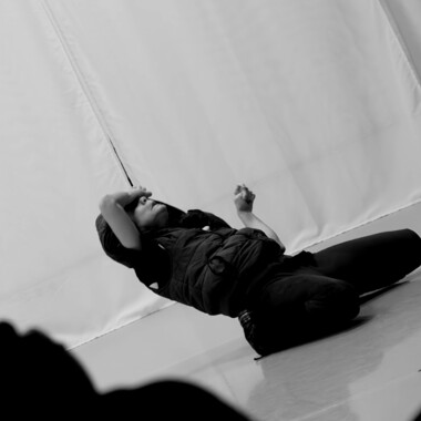 Ein schwarz/weiß Foto. Eine Person kniet auf dem Boden eines hellen Trainingsraumes und lehnt sich zurück. Die eine Hand liegt auf der Stirn, der andere Arm ist gebeugt. Die Haltung wirkt dramatisch oder performativ.