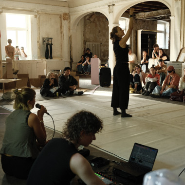 Eine Person steht auf einer hellen Tanzfläche mitten in einem großen Raum. Sie hebt einen Arme nach oben. Rundherum sitzen zahlreiche Zuschauer:innen am Boden und beobachten die Szene. Im Vordergrund sitzen zwei Personen, eine Person bedient technische Geräte, die andere spricht in ein einem Mikrofon.