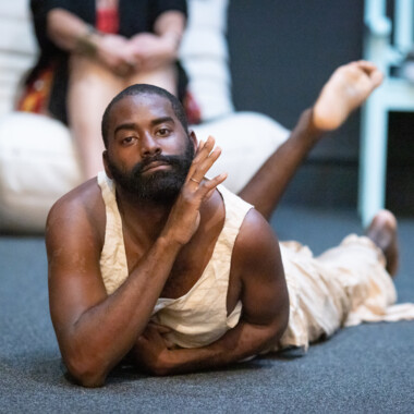 Eine Szene aus LAX. Eine Nahaufnahme von Jerron, ein Schwarzer Mann, der bäuchlings auf einem Boden liegt und direkt in die Kamera schaut. Der Oberkörper ist leicht gehoben und einen Arm so angewinkelt, dass die Hand nahe am Gesicht ist.