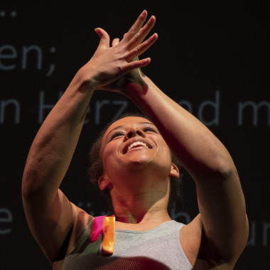 Naomi Sanfo-Ansorge applauding. Naomi wears a gray tank top and smiles. She holds her hands up as she applauds. Behind her is a black wall on which words can be seen in blue letters.