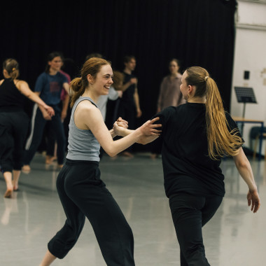 Ein Foto aus einem Workshop von Cara Rother. Es zeigt mehrere Menschen in einem Raum, die miteinander tanzen und sportlich gekleidet sind. Im Bildvordergrund Cara Rother. Sie lächelt die Person vor ihr an, dessen Hand sie mit ihrer linken Hand hält. Ihre rechte Hand liegt auf dem Oberarm dieser Person, die von hinten zu sehen ist.
