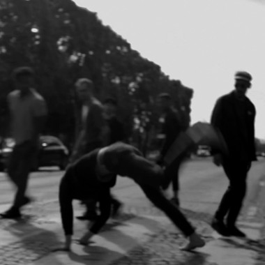 Black and white photo of a situation on a street with cobblestones, trees and cars in the background. Nine people in everyday clothes circle another person who appears to be doing a handstand. They are all on a crosswalk.