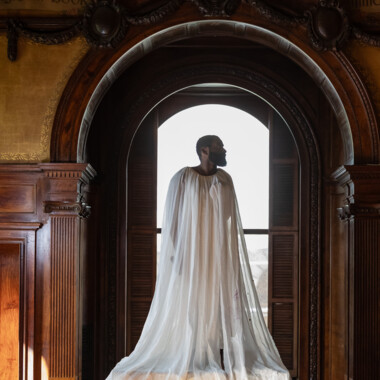 Ein Foto von Jerron. Er steht erhöht auf einem großen, mit Stoff bedeckten Objekt vor einem hohen Fensterbogen in einem holzgetäfelten Raum. Ein transparenter, bodenlanger Stoff fällt von den Schultern der Person herab.