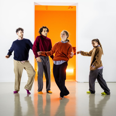 The four performers Yuri Fortini, Talal Mouzanar, Nina Melcher and Vivien Kaluza stand in a row in front of a white wall, each dancing on their own. They ask colourful socks, plain jumpers in blue, red, orange and brown. An illuminated room with orange-coloured walls can be seen in the background.