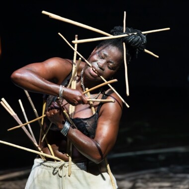 Eine Szene aus Épique ! (pour Yikakou). Eine Person steht auf einer Bühne, sie trägt einen schwarzen BH und eine helle Hose. Der Oberkörper ist nach links geneigt und sie ist von vielen Stäben umgeben. Sie hält einige davon mit Armen und Mund fest, andere Stecken in den Haaren und dem Kostüm. Der Hintergrund ist dunkel.