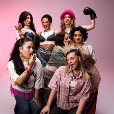 Sieben Personen posieren vor einem rosa Hintergrund. Sie stehen und sitzen eng beieinander und zeigen verspielte oder ausdrucksstarke Posen. Einige tragen auffällige Kleidung, Accessoires oder Make-up. Eine Person trägt einen Boxhandschuh, den sie in die Luft streckt.