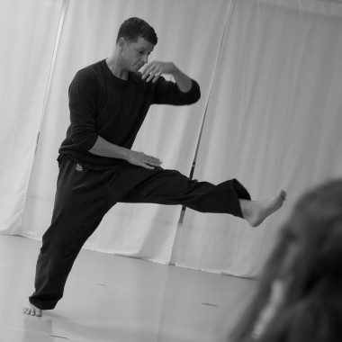 Ein schwarz/weiß Foto. Eine Person bewegt sich barfuß in einem hellen Trainingsraum. Ein Bein ist gestreckt nach vorne angehoben, ein Arm vor das Gesicht geführt. Die Haltung wirkt tänzerisch oder kampfsportähnlich.