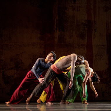 Eine Szene aus 2x4 von A.I.M by Kyle Abraham. Vier Tänzer:innen in bunten Kostümen stehen eng zusammen. Alle beugen ihre Oberkörper nach links und lehnen sich teils aneinander und übereinander. Ihre Körper bilden gemeinsam eine annähernd runde Form.