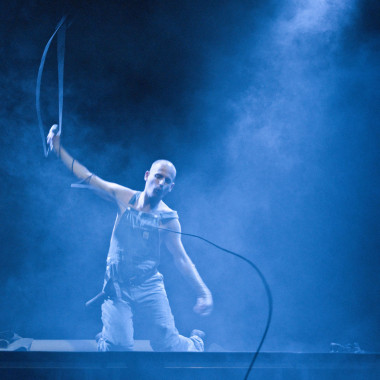 A scene from Work Body: The photo shows Michael Turinsky sitting on stage in dungarees. He is holding his right arm in the air and a wired microphone in his hand. The microphone cable swings in the air.