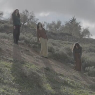 The photo shows three people standing on a vegetated hillside. They are wearing loose clothing in earthy tones and are looking in different directions. The sky is cloudy, and trees and shrubs can be seen in the background.