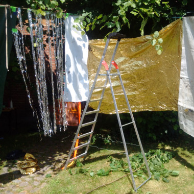 Mehrere Stoffbahnen hängen im Freien an einer Leine, darunter goldene Folie, ein weißes Tuch und silberglänzende Streifen, die eine Halbkreis bilden. Davor in der Mitte steht eine Aluminiumleiter auf einer Wiese.