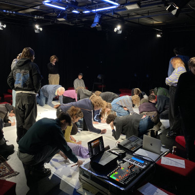 Viele Personen knien oder hocken dicht beieinander auf einer weißen Bodenfläche in einem dunklen Bühnenraum. Sie schreiben etwas auf Zettel und Plakate, die auf dem Boden liegen. Links steht ein Tisch mit Audio-Equipment, Mischpult und Laptop.
