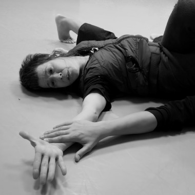 Ein schwarz/weiß Foto. Zwei Personen liegen auf dem Boden in einem hellen Trainingsraum. Im Fokus des Bildes sind zwei Hände, die sich berühren. Eine Person liegt ausgestreckt auf dem Rücken, den Blick auf die Hände gerichtet. Von der anderen Person ist nur der ausgestreckte Arm im Bild.