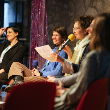 Podium-Situation: Auf dem Podium sind von links nach rechts Mónica García Vicente, Selina Glockner, und Heike Bröckerhoff zu sehen. Sie lachen, während Heike ins Publikum spricht.