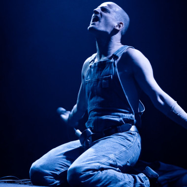 A scene from Work Body: The photo shows Michael Turinsky sitting on stage in dungarees. He stretches his arms back and throws his head back. He seems to be shouting something. The stage is bathed in blue light.