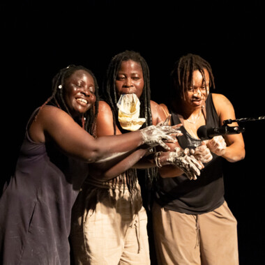 Eine Szene aus Épique ! (pour Yikakou).Drei Personen stehen dicht nebeneinander auf einer Bühne. Ihre Hände sind mit einer weißen Substanz beschmiert. Die Person in der Mitte hält eine Tüte im Mund. Alle schauen konzentriert auf ein Mikrofon vor ihnen.