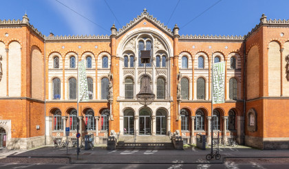 Frontalansicht auf das Künstlerhaus Hannover. Es ist im Rundbogenstil gebaut und mit vielen Fenstern, Figuren und Reliefs verziert. Vor dem Gebäude hängt ein großer Kronleuchter über der Straße.