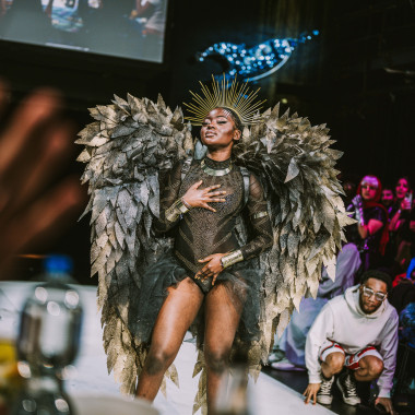 Catwalk situation: A person with a black and gold bodysuit, gold crown and large gold-coloured wings presents herself on the catwalk. They has their eyes closed and has placed their hands on their body. Many other people are standing around them, clapping and cheering them on.