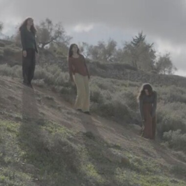Das Foto zeigt drei Personen, die an einem bewachsenen Hang stehen. Sie tragen lockere Kleidung in Erdtönen und blicken jeweils in unterschiedliche Richtungen. Der Himmel ist bewölkt, im Hintergrund sind Bäume und Sträucher.