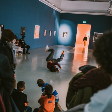 Ein langer Rad mit blauen Wänden, Publikum an den Seiten des Raumes. In der Mitte Talal Mouzanar in einer Breakdance-Bewegung auf dem Boden. In der Ferne steht Yuri Fortini im Türrahmen zum nächsten Raum und schaut zu Talal.