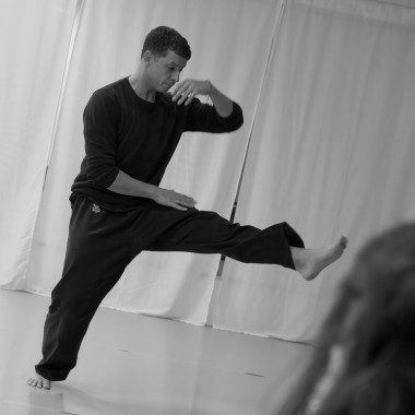 A black-and-white photograph. A person is moving barefoot in a bright training room. One leg is stretched out in front, one arm is raised in front of the face. The posture appears dance-like or martial arts-like.