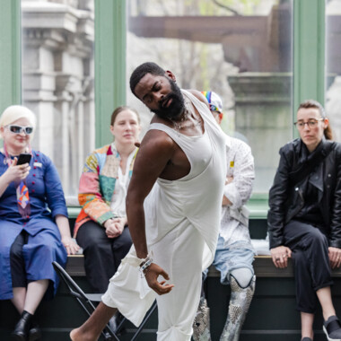 Eine Szene aus LAX. Jerron tanzt in einem weißen Kostüm. Der Körper ist zur Seite geneigt, der Blick über die Schulter. Hinter ihm sitzt Publikum auf einer Bank und schaut ihm zu.