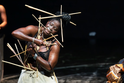 Eine Szene aus Épique ! (pour Yikakou). Eine Person steht auf einer Bühne, sie trägt einen schwarzen BH und eine helle Hose. Der Oberkörper ist nach links geneigt und sie ist von vielen Stäben umgeben. Sie hält einige davon mit Armen und Mund fest, andere Stecken in den Haaren und dem Kostüm. Der Hintergrund ist dunkel.