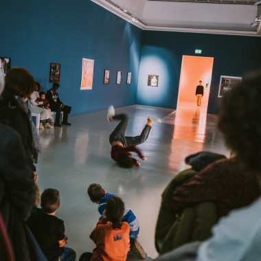 A long wheel with blue walls, audience on the sides of the room. In the centre, Talal Mouzanar is breakdancing on the floor. In the distance, Yuri Fortini stands in the doorway to the next room and looks at Talal.