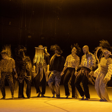 Eine Gruppe von sieben Performer*innen aus dem Stück TARAB auf einer Bühne, die in gelbes Licht eingetaucht ist. Sie stehen in einer Reihe und halten sich dabei an den Händen. Sie tragen weite Kleidung in Erdtönen und bewegen sich gleichzeitig. Dabei werfen sie ihre Haare in die Luft. 