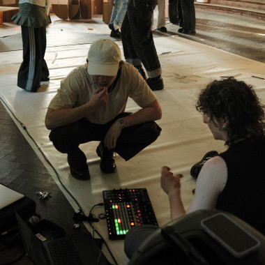Zwei Personen auf einer hellen Bodenfläche in einem großen, leicht abgenutzt wirkendem Raum. Sie sitzen vor einem kleinen Mischpult und scheinen sich darüber zu unterhalten.