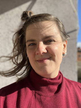 Ein Portrait von Anna Wieczorek: Anna trägt einen roten Rollkragenpulli und lächelt. Sie hat blaue Augen und lange, hellbraune Haare, die sie am Oberkopf zusammengebunden hat. Sie trägt silberne Ohrringe. 
