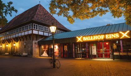 The Ballhof Eins building with a sign reading Ballhof Eins. The yellow lettering is brightly illuminated. The sign is located above the entrance, which is glazed. There is a lantern in front of the building, which is also illuminated.