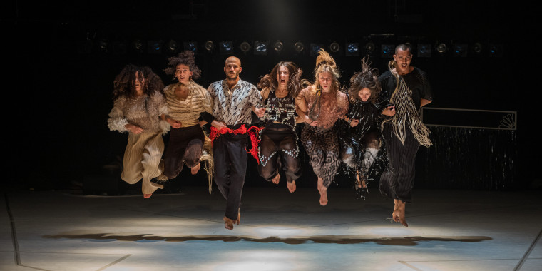 A group of seven people have hooked each other in a row and are floating in the air after a jump. They wear loose clothing in earthy colours and look at the camera with strong expressions on their faces.