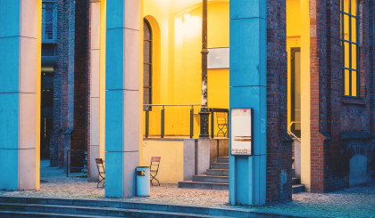 Exterior view of Cumberland. The high entrance area with stone arches is painted yellow and lined with stone and iron pillars. Eight steps lead to a level in front of the entrance door, which is also used as a smoking area. There are a few folding chairs in the entrance area.