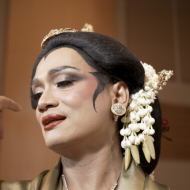 Ein Porträt von Rianto. Rianto ist auffällig geschminkt und trägt eine goldene Haarverzierung und weißen Blütenschmuck im Haar. Der Gesichtsausdruck ist ruhig und konzentriert auf die Hand gerichtet, die elegant erhoben ist.