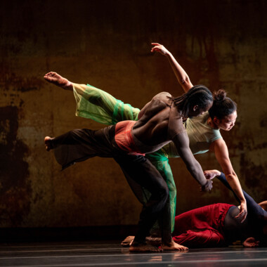 Eine Szene aus 2x4 von A.I.M by Kyle Abraham. Zwei Tänzer:innen in bunten Kostümen bewegen sich zusammen. Sie sind mit dem Rücken zum Publikum ausgerichtet. Eine Person hebt ein Bein hoch, streckt die Arme auseinander und lehnt sich nach vorne, während eine andere Person sie seitlich stützt.