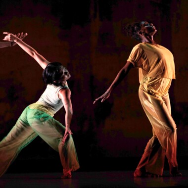 Eine Szene aus 2x4 von A.I.M by Kyle Abraham. Zwei Tänzer:innen bewegen sich auf einer Bühne. Eine Person trägt ein gelbes Kostüm, sie beugt ihren Oberkörper nach hinten und streckt einen Arm nach hinten aus. Die andere Person ist grün gekleidet, sie steht in einem Ausfallschritt, beugt sich nach vorne und streckt die Arme auseinander.