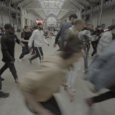 A high room that looks like an empty factory hall. A group of about 15 people move in a crowd from left to right through the picture, they seem to be running. They are all wearing different clothes and trainers. One person on the right of the picture appears to be breakdancing, the eyes of the other people are on the person.