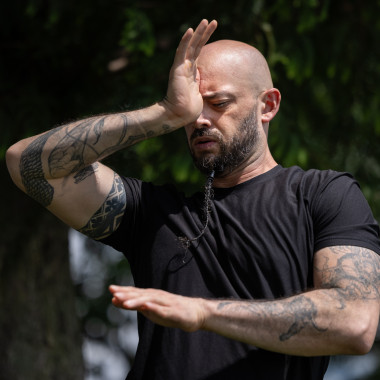 Christopher atmet tief ein, schließt die Augen und berührt mit seiner rechten Hand sein drittes Auge, wodurch er eine Längsebene markiert. Sein linker Arm zeichnet einen Kreis in der Querfläche. Er trägt ein schwarzes T-Shirt, das seine Arme freilegt, die mit mehreren Tätowierungen verziert sind.