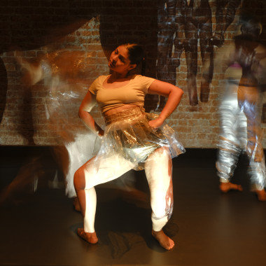 A stage situation: three dancers from the piece "Hallucinations of an Artifact", behind them a brick wall on which the sculpture Dancing Girl is projected several times side by side. The performer in the foreground has tied her dark hair into a bun and is lying sideways on the floor. She is leaning on the floor with her hands spread wide apart and looking down. The person in the centre is leaning on the floor with their feet and hands, with the middle part of their body in the air. The third person in the background lies sideways on the floor and looks at the projection behind them. As with the projection of the dancing girl, one hand rests on the hip.