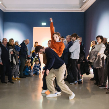 Vivien Kaluza, Yuri Fortini and Nina Melcher dance together. There is an audience around them, some children are sitting on the floor. The walls in the background are dark blue.
