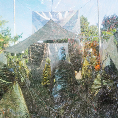 Blick durch eine durchsichtige, knitterige Folie auf eine Installation im Freien. Unter einem Zeltdach stehen mehrere gold- und silberfarbene, figurähnliche Formen. Bäume und Himmel sind im Hintergrund sichtbar.