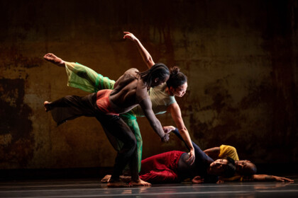 Eine Szene aus 2x4 von A.I.M by Kyle Abraham. Zwei Tänzer:innen in bunten Kostümen bewegen sich zusammen. Sie sind mit dem Rücken zum Publikum ausgerichtet. Eine Person hebt ein Bein hoch, streckt die Arme auseinander und lehnt sich nach vorne, während eine andere Person sie seitlich stützt. Zwei weiter Personen liegen daneben auf dem Boden.