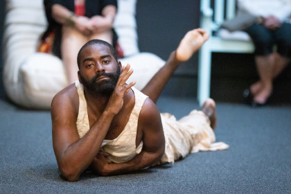 Eine Szene aus LAX. Eine Nahaufnahme von Jerron, ein Schwarzer Mann, der bäuchlings auf einem Boden liegt und direkt in die Kamera schaut. Der Oberkörper ist leicht gehoben und einen Arm so angewinkelt, dass die Hand nahe am Gesicht ist.