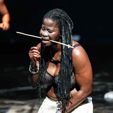 Eine Szene aus Épique ! (pour Yikakou). Eine Person steht auf einer Bühne und hält einen langen Stock zwischen den Zähnen. Sie trägt ein schwarzen BH und eine helle Hose. Der Hintergrund dunkel.