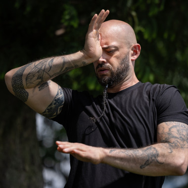 Christopher takes a deep breath with his eyes closed while his right hand touches his third eye, marking a longitudinal plane. His left arm draws a circle in the transverse plane. He is wearing a black t-shirt that exposes his arms, which are decorated with multiple tattoos.