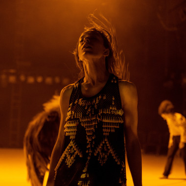 A performer from the piece "TARAB". The person stands with closed eyes and slightly open mouth on a stage bathed in orange-coloured light. He*she throws his*her head back slightly, the long hair is in motion. The performer is wearing a sleeveless dark top with decorative curtain hooks. Two other performers can be seen moving in the background. 