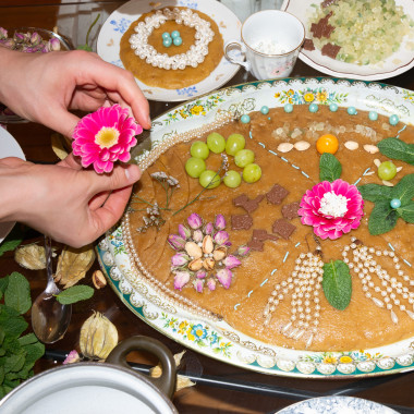 A large plate of halva. The dessert is lavishly decorated with fruit, flowers and nuts. Next to it are smaller plates with other dishes and in the left half of the picture two hands holding a flower.