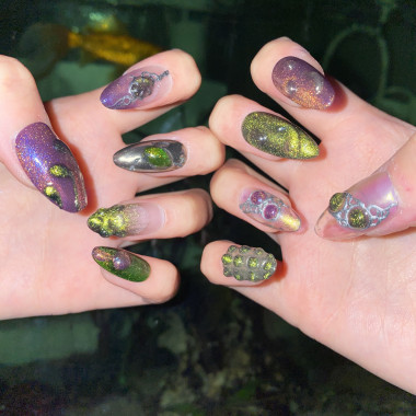 Zwei Hände, die ein Nageldesign von Nail Artistin Hong Ngoc Le zeigen. Die Nägel sind oval und lang. Sie schimmern lila und weisen grün schimmernde Akzente in Form von organischen Formen auf.